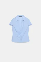 Camisa de color celeste con cuello solapa, escote en pico y manga corta. Presenta un diseño entallado con detalle de frunces verticales en la parte central y cierre frontal con botones.