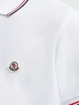 Polo de piqué de algodón en color blanco, con cuello y puños acanalados que presentan detalles de rayas tricolor. Cuenta con cierre frontal de botones y el logo de la marca aplicado en el pecho.