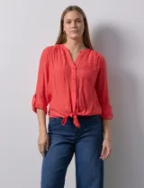 Blusa blanca de gasa marca Zac & Rachel, con cuello a la base, escote en V, abertura central con botones, bolsillo aplicado en el pecho y mangas largas regulables con presilla y botón. Tiene terminación opcional de nudo en el ruedo.