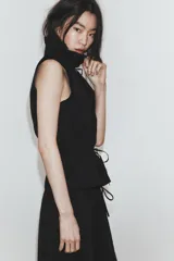 Chaleco negro de diseño fluido con cuello subido y cierre frontal de cremallera metálica. Presenta cordones ajustables con lazadas en el cuello y en la cintura.