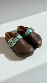 Sandalias de cuero marrón de la línea Ottenta de Carolina de Cunto, con diseño estilo zueco, tira ajustable con hebilla metálica y detalles pintados a mano en tonos turquesa y dorado.