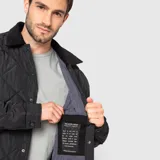 Chaqueta acolchada de diseño romboidal en color negro, con cuello camisero de textura aterciopelada y cierre frontal mediante botones a presión.