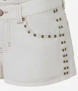 Short de sarga color blanco roto, modelo micro, con trabillas en la cintura, cierre tradicional, bolsillos y tachuelas aplicadas desde los bolsillos hasta los laterales. Presenta costura aparente en contraste.
