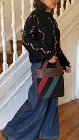 Bolso tipo matera confeccionado en cuero vacuno, con diseño estructurado y manija superior integrada. Presenta un cuerpo con franjas diagonales en gamuza de colores contrastantes (amarillo, verde y azul) sobre una base de cuero marrón, con detalles de costuras artesanales en los bordes.