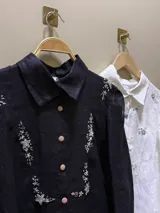 Camisa de lino con cuello clásico y cierre frontal de botones. Presenta bordados artesanales detallados con motivos florales y estrellas en la parte frontal y hombros.