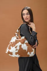 Bolso tipo hobo de gran capacidad con estampado animal print de vaca en tonos marrón y blanco. Presenta una etiqueta de cuero con el logo de la marca en el frente y un diseño de caída relajada.