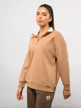 Buzo de felpa color tostado con cuello alto y medio cierre frontal. Presenta un diseño de corte holgado con hombros caídos, puños y ruedo acanalados, y el logo de la marca bordado en el pecho.