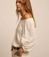 Blusa blanca con lunares dorados, escote hombro a hombro y mangas largas abullonadas con ajuste en los puños.