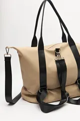 Bolso tipo tote color beige con detalles en negro, confeccionado en material sintético liso. Cuenta con doble asa superior, correa ajustable y extraíble, cierre principal superior y un compartimento inferior independiente con cierre. Presenta correas decorativas verticales con hebillas metálicas y logo grabado en la base.