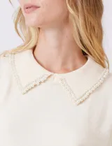 Buzo tejido color beige, marca Nanette, con cuello a la base y solapa decorada con apliques de perlas. Tiene manga corta con puño y ruedo acanalado.