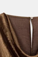 Blusa de manga larga con cuello fluido tipo drapeado y abertura en los puños. Presenta un acabado satinado con textura y cierre posterior con cremallera oculta.