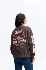 Buzo de algodón color marrón chocolate, con cuello redondo y diseño holgado. Presenta una estampa gráfica en la espalda con motivos de atardecer y montañas, junto con texto decorativo. Además, incluye una estampa en la manga con letras grandes.