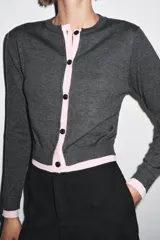 Chaqueta de punto gris de manga larga, cuello redondo y cierre frontal con botones oscuros. Presenta ribetes y detalles en color azul eléctrico en el frente, puños y ruedo.