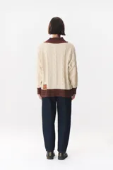 Buzo de tejido de punto con diseño de trenzas, color crema con detalles en marrón en cuello, puños y ruedo. Presenta líneas finas en contraste y pequeñas costuras decorativas en forma de cruz.