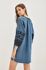 Vestido corto de tela tipo denim con bordados geométricos en color negro en el pecho y las mangas. Presenta escote en V, mangas tres cuartos con puños elásticos y un corte holgado.