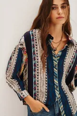 Camisa de manga larga con estampado étnico multicolor, cuello clásico y cierre frontal con botones. Presenta un diseño de rayas verticales con patrones geométricos y detalles en tonos azules, rojos, amarillos y beige.