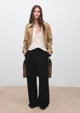 Trench coat color beige, con cuello de solapa, diseño cruzado y cierre frontal de botones. Presenta trabillas en los puños, cinturón ajustable y bolsillos laterales.
