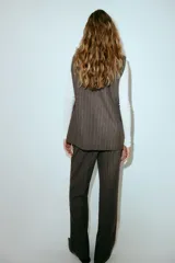 Pantalón sastrero de tiro alto, color marrón oscuro con finas rayas verticales blancas. Presenta cierre frontal con botón oculto y corchetes, bolsillos al bies y bolsillos decorativos traseros. Las piernas son rectas y tienen pliegues marcados al frente y atrás.