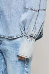 Camisa de denim celeste con calce regular, cuello camisero, cierre frontal con botones metálicos y mangas estilo balloon con puños largos ajustables.