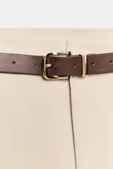 Pantalón tobillero de tiro medio, color beige, con pinzas delanteras y bolsillos frontales. Incluye un cinturón fino de cuero marrón con hebilla metálica dorada y cierre frontal con cremallera, botón interior y gancho.