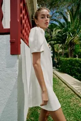 Vestido corto de color blanco con cuello redondo y mangas cortas abullonadas. Presenta un cierre frontal con botones decorativos de colores.