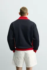 Chaqueta deportiva de corte regular, confeccionada en tejido piqué con interior forrado en red. Presenta un diseño color block en negro y rojo con detalles en blanco, cuello subido, cierre frontal con cremallera y bolsillos laterales con cierre.