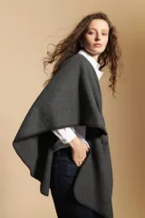 Poncho de tejido suave color beige con diseño envolvente y terminaciones con costuras decorativas en contraste color negro. Presenta un detalle de lazo frontal integrado.