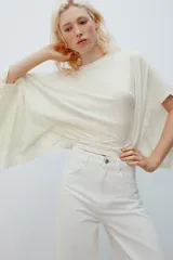 Camiseta tipo capa oversize de color blanco, confeccionada en hilatura de algodón 100%. Presenta cuello redondo, manga corta y un diseño de bajo asimétrico con caída pronunciada en uno de los laterales.