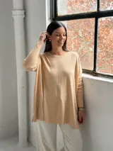Sweater de punto color beige, de corte holgado y diseño asimétrico con tajos laterales. Presenta cuello redondo y mangas largas con puños acanalados, además de un detalle de costura vertical central.