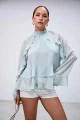 Blusa verde menta de gasa, con cuello alto fruncido, mangas largas amplias y detalles de volados en cascada en el frente y las mangas.