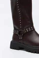 Bota de caña media estilo biker, confeccionada en cuero sintético color marrón oscuro. Presenta una suela track robusta, detalle de tachas metálicas verticales a lo largo de la caña y un arnés decorativo con argolla metálica en el tobillo.