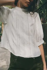 Blusa blanca de cuello alto con detalles de puntilla, manga corta abullonada y cierre posterior con botón.