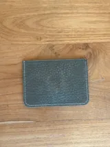 Tarjetero de cuero color coral con acabado brillante, diseño de ranuras en V y placa metálica con el logo de la marca en el frente.