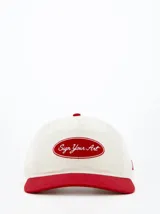 Gorra de béisbol color crema con visera y botón superior rojos. Presenta un logo bordado en el frente con la frase "Sign Your Art" en rojo.