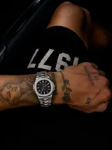 Reloj de pulsera con caja de acero inoxidable de 40mm y esfera negra con textura horizontal. Presenta marcadores de hora luminiscentes, ventana de fecha a las tres y brazalete de acero inoxidable con acabado cepillado y pulido.