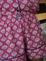 Pijama de dos piezas, compuesto por una camisa de manga corta con cuello solapa y botones frontales, y un short a juego. El conjunto es de color azul claro con un estampado repetitivo de pequeños motivos florales en color rosado/rojo, y ribetes de color rojo en los bordes de la camisa y el short.