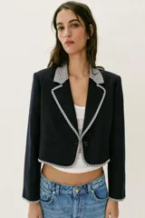 Blazer corta de color negro con cuello solapa, manga larga y hombreras. Presenta detalles combinados con estampado de cuadro vichy en el cuello, los bordes de la solapa, el ruedo y los puños. Cuenta con cierre frontal de un botón y falsos bolsillos delanteros de vivo.