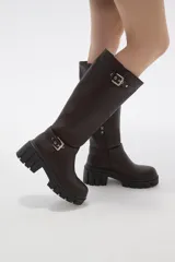 Bota de caña alta color negro, confeccionada en eco cuero, con suela track de goma y detalle de hebilla metálica en el tobillo y en la parte superior de la caña.