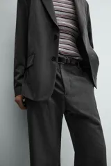 Pantalón de vestir gris oscuro, corte ancho (flare fit) y tiro alto, con bolsillos laterales y falsos bolsillos de vivo en la parte trasera. Presenta un cierre frontal con cremallera y gancho oculto, y botones a presión ocultos en el bajo.