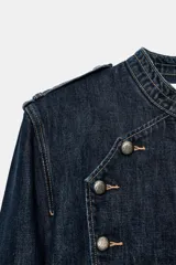 Chaqueta de denim de corte corto, color azul oscuro, con cuello redondo, hombreras y cierre frontal con cremallera. Presenta diseño de inspiración militar con doble botonadura metálica decorativa en el frente, trabillas con botón en los hombros y bajo elástico.