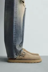 Zueco tipo mule de hombre, color beige topo, fabricado en piel con acabado serraje. Presenta una tira con hebilla metálica cuadrada en el empeine y suela gruesa de goma en color natural contrastante.