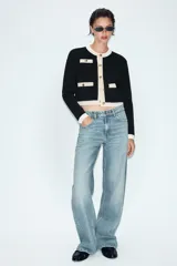 Chaqueta de punto de corte recto y manga larga, con cuello redondo. Presenta un diseño bicolor en negro y crema, con cierre frontal de botones dorados y bolsillos de plastrón en el delantero con detalles en contraste.