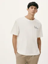 Camiseta blanca de algodón con cuello redondo y mangas cortas. Estampa rectangular con borde negro que contiene la frase "NATURAL ATMOSPHERE" en letras mayúsculas y una imagen de montañas y mar.
