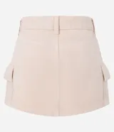 Pollera corta de sarga color beige claro, con cintura con trabillas y cierre de botón y cremallera. Presenta bolsillos cargo funcionales a los lados.