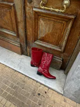 Bota estilo biker de caña alta, confeccionada en cuero vacuno color rojo cherry. Presenta un diseño con múltiples tiras horizontales con hebillas metálicas, punta redondeada y taco cuadrado bajo de madera.