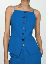 Chaleco entallado de color azul, con cuello halter, tirantes finos y cierre frontal con botones oscuros. Presenta dos bolsillos de ribete delanteros y forro interior.