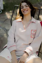 Buzo color crema con cuello polo, mangas largas y puños ajustados. Presenta un bolsillo en el pecho con bordados de frutas y la inscripción "Bom Faro by Coppelia". Los bordes del cuello, el bolsillo y los puños están decorados con puntadas rojas.