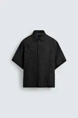 Camisa negra de manga corta con cuello solapa y textura arrugada o plisada. Corte relajado (relaxed fit) con aberturas laterales y cierre frontal de botones.