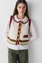 Chaleco de punto estilo lambswool color crema, con cuello estilo bebé y bordes festoneados en color bordó. Presenta cierre frontal con botones, bolsillo pequeño en el frente y detalles de rayas en la parte inferior en tonos bordó, verde oliva y mostaza.