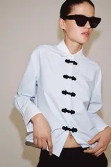 Camisa de algodón con cuello solapa y puños en color blanco a contraste. Presenta cierre frontal con alamares negros y bajo asimétrico redondeado.